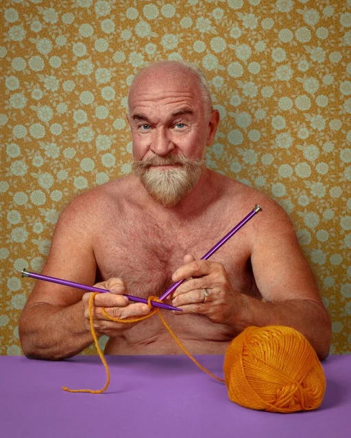 Middle-aged man with beard and bare chest knitting orange yarn with purple knitting needles indoors against a floral wallpaper background.